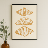 Framed artwork of croissants on a wall above a white cabinet with a vase and books.